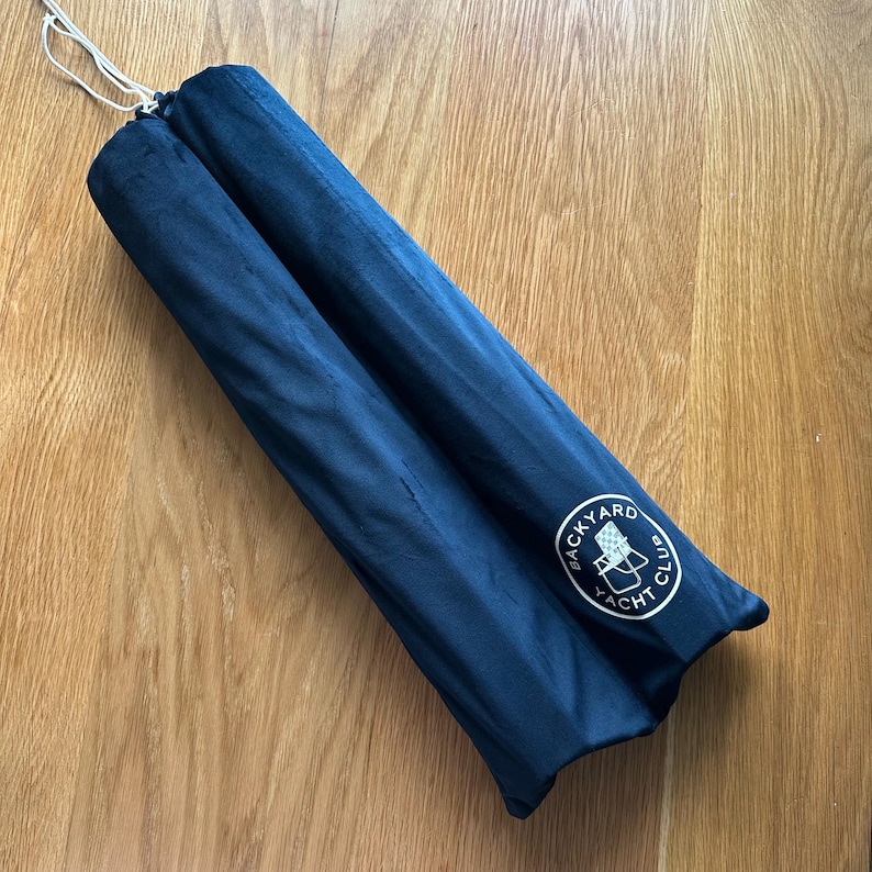 May include: A navy blue, cylindrical item, possibly a rolled-up umbrella or similar product, is shown. The item is secured with a white drawstring. The Backyard Yacht Club logo is visible on the side.