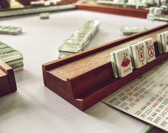 May include: A wooden mahjong tile rack holding white tiles with red and green symbols, including a tile marked SOUTH and a red fan icon. A printed score card with numerical sequences and the year 2025 is placed on the light-colored playing surface.