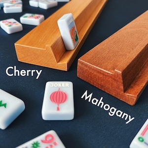 May include: Cherry and mahogany wooden mahjong tile holders with white tiles. The tiles have colorful designs and text. One tile is standing upright, displaying a flower. The word "Cherry" is written on the left, and "Mahogany" on the right.