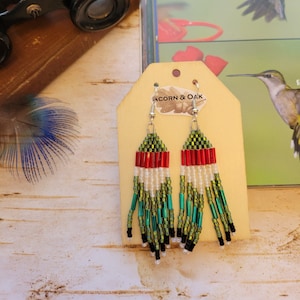 May include: Handmade beaded earrings with a fringe design. The earrings feature a pattern of red, white, green, and black beads. The earrings are hanging from a wooden tag that says "ACORN & OAK".