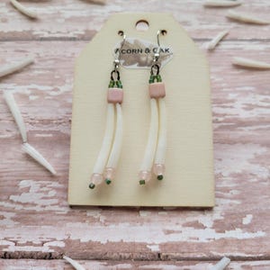 May include: Handmade earrings with a unique design. Each earring features a pale pink square bead, white strands, and small green and pink beads. The earrings hang from silver hooks and are displayed on a wooden tag with the text "CORN & CAK".