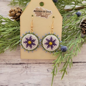 May include: Handmade beaded earrings with a circular design. The earrings feature a white background with a purple and yellow floral pattern, surrounded by green and blue beads. The earrings hang from gold-colored hooks. The earrings are displayed on a wooden tag that reads "ACORN & OAK".