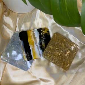 May include: Three coasters with different designs. One coaster is silver with glitter, one is black and white with gold accents, and one is gold with glitter.