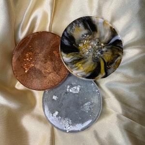 May include: Three round resin coasters with a unique, abstract design. The coasters feature a black, white, and gold swirl pattern, a copper-colored swirl pattern, and a gray swirl pattern with silver flakes.