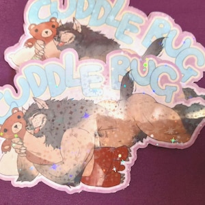 Puede incluir: Dos pegatinas rosas y azules con el texto "Cuddle Rug" e ilustraciones de una criatura peluda abrazando a un osito.