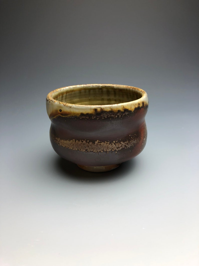 May include: A small, brown ceramic bowl with a white rim and a speckled glaze. The bowl has a rounded shape and a slightly flared lip.