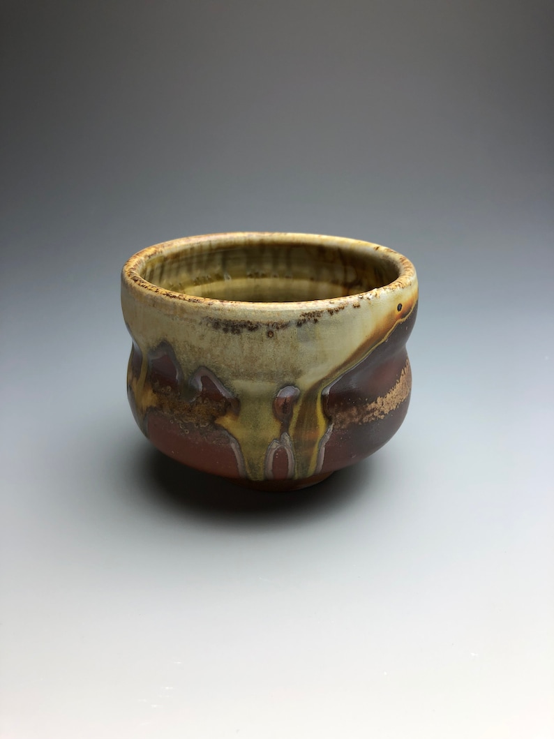 May include: A small, brown ceramic bowl with a textured, drippy glaze. The bowl has a rounded shape and a slightly flared rim.