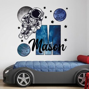 Könnte beinhalten: Ein Wandaufkleber mit einem Cartoon-Astronauten, der neben einem grauen Mond und zwei blauen Planeten schwebt. Ein großer Buchstabe M mit einem blauen Nebelmuster befindet sich hinter dem Namen Mason in schwarzer Schreibschrift.