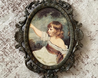 May include: An oval wall frame made of dark bronze-toned metal with ornate scrollwork and fleur-de-lis accents. The frame contains a portrait of a child with shoulder-length brown hair, wearing a white garment with a dark sash, pointing to the left.