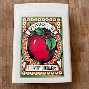 Puede incluir: Caja rectangular blanca con una manzana roja y hojas verdes en la parte delantera. La caja tiene un borde dorado y verde con el texto "AVON COUNTRY ORCHARDS" en una pancarta.