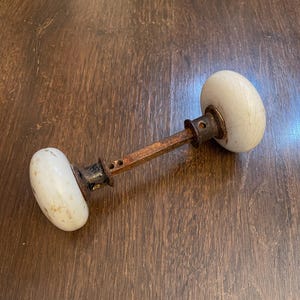 May include: An antique door knob with two oval, off-white ceramic handles connected by a rusty metal rod. The metal shows signs of age and wear, with dark spots and discoloration. The knob is set against a brown wooden surface.