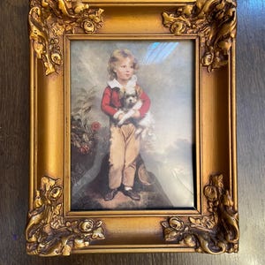 May include: A framed portrait featuring a young person in a red jacket and tan pants holding a small dog. The gold-colored frame has ornate floral and scrollwork details. The artwork has a vintage aesthetic.