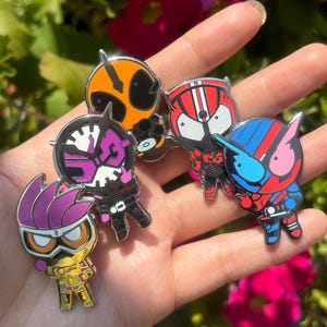 Könnte beinhalten: Eine Sammlung von fünf farbenfrohen Emaille-Anstecknadeln mit stilisierten Charakteren mit einzigartigen Helmdesigns. Die Anstecknadeln werden in einer Hand vor dem Hintergrund leuchtend rosa Blüten und grünem Laub gehalten.