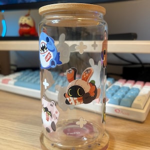 May include: A clear glass jar with a bamboo lid and various cartoon character stickers. The stickers feature a blue shark, a brown bunny, a white cat, and a brown character with a red scarf. The jar is sitting on a wooden surface.