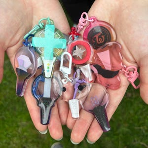 Puede incluir: Una colección de llaveros transparentes y coloridos con varios diseños, incluyendo una cruz, una estrella y un logotipo. Los llaveros se sostienen en manos abiertas sobre un fondo verde.