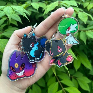 May include: Four colourful cartoon character keychains. The keychains are made of clear acrylic with a holographic finish. The characters are a purple and red ghost, a black and blue dragon, a black and red creature with a skull face, and a green and white plant-like creature.