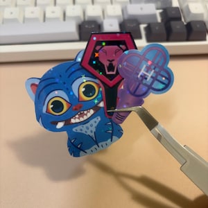 May include: A blue cartoon tiger sticker with large yellow eyes and a toothy grin. A red and black shield-shaped sticker with a lion graphic and a purple clover-shaped sticker are also visible. The stickers are being held by tweezers.