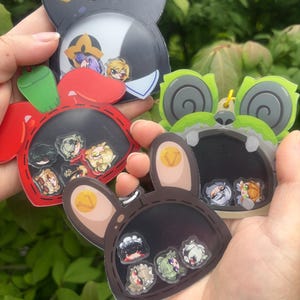 May include: A collection of colorful, cartoon-style character keychains. The keychains are shaped like animal heads, including a black and gold bunny, a red bunny, a brown bunny, and a green frog. Each keychain has a clear window with small character figures inside.