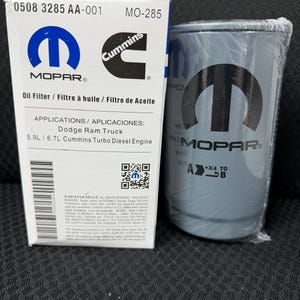 Puede incluir: Un filtro de aceite Mopar y su caja. La caja es blanca con texto azul y negro, incluyendo los logotipos de Mopar y Cummins. El filtro es gris con el logotipo de Mopar y est&aacute; envuelto en pl&aacute;stico transparente. La caja dice "Filtro de aceite" y "Dodge Ram Truck".