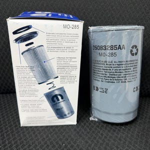 Puede incluir: Se muestran dos filtros de aceite automotrices, uno en caja y otro envuelto en pl&aacute;stico. La caja es blanca y azul, con texto y diagramas. El filtro envuelto es gris con texto impreso sobre un fondo oscuro.
