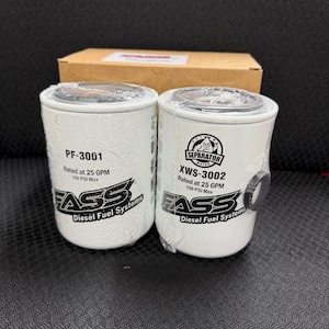 May include: Two white cylindrical fuel filters with black text and logos, labeled PF-3001 and XWS-3002. The filters are from FASS Diesel Fuel Systems and are rated at 25 GPM and 100 PSI Max. A cardboard box is in the background.