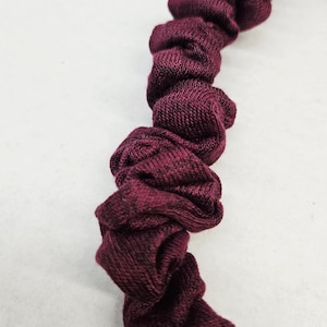 Puede incluir: Primer plano de una goma para el pelo burdeos. La tela est&aacute; fruncida y retorcida, creando una apariencia texturizada y con volantes. La goma para el pelo est&aacute; hecha de un material suave y tejido. Este accesorio para el cabello es una adici&oacute;n elegante a cualquier peinado.