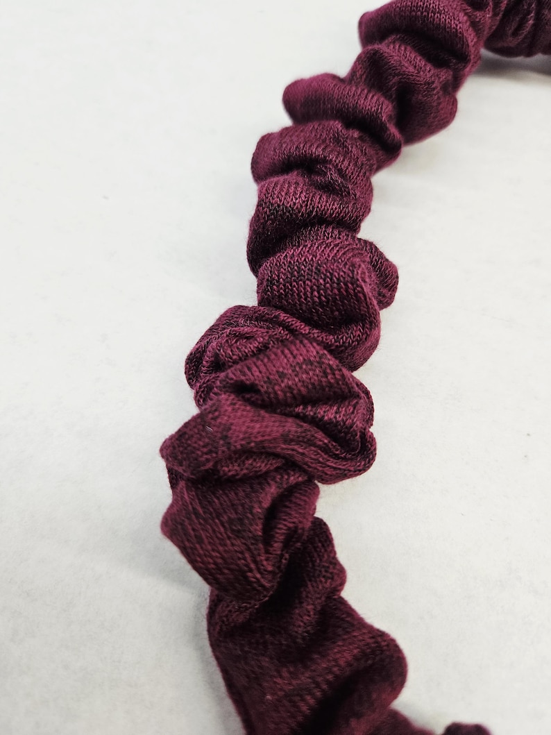 Puede incluir: Primer plano de una goma para el pelo burdeos. La tela est&aacute; fruncida y retorcida, creando una apariencia texturizada y con volantes. La goma para el pelo est&aacute; hecha de un material suave y tejido. Este accesorio para el cabello es una adici&oacute;n elegante a cualquier peinado.