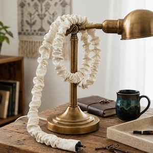 May include: A brass desk lamp with a rounded head and a sturdy base, featuring a long electrical cord encased in a gathered, off-white fabric sleeve that loops around the lamp's central vertical post, ending in a standard two-prong plug.