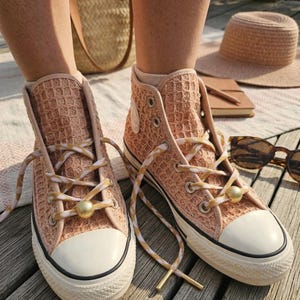 Könnte beinhalten: Pfirsichfarbene High-Top-Sneaker mit Waffelstruktur. Sie haben weiße Gummisohlen, passende Schnürsenkel mit goldenen Akzenten und goldene Schnürsenkelperlen. Ein Strohhut und eine Sonnenbrille sind im Hintergrund zu sehen.