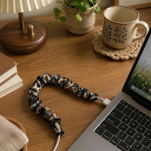 Può includere: Un proteggi cavo nero, bianco e beige è collegato a un laptop. Il proteggi cavo è realizzato in tessuto strutturato. Sono visibili anche una scrivania in legno, una lampada, una tazza e una piccola pianta.