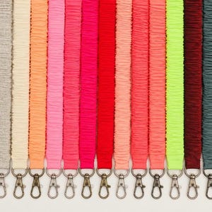 May include: A collection of nineteen fabric straps arranged vertically in a row, each featuring a gathered, ruched texture and a metal lobster clasp at the bottom. The straps are displayed in a variety of solid colors including black, yellow, gray, pink, red, orange, neon green, teal, and beige.