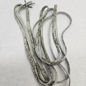 Puede incluir: Un juego de cordones grises claros con un estampado floral repetido en blanco y gris oscuro. Los cordones est&aacute;n dispuestos en un patr&oacute;n suelto y en bucle sobre un fondo blanco. Los extremos de los cordones est&aacute;n anudados.