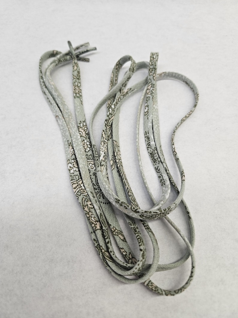 Puede incluir: Un juego de cordones grises claros con un estampado floral repetido en blanco y gris oscuro. Los cordones est&aacute;n dispuestos en un patr&oacute;n suelto y en bucle sobre un fondo blanco. Los extremos de los cordones est&aacute;n anudados.
