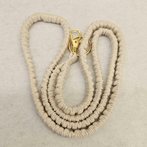 May include: A beige macrame lanyard featuring a textured, knotted cord design. The lanyard includes a gold-toned metal swivel lobster clasp and a circular split ring attached at the top, providing a functional connection point for various items.