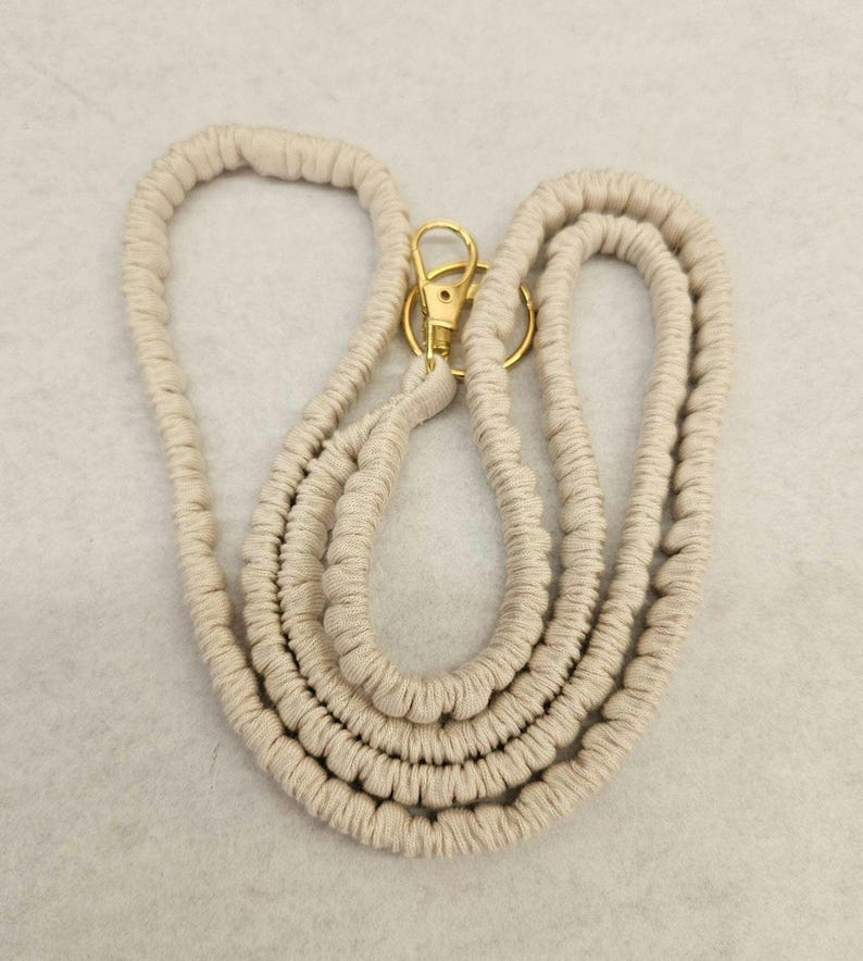 May include: A beige macrame lanyard featuring a textured, knotted cord design. The lanyard includes a gold-toned metal swivel lobster clasp and a circular split ring attached at the top, providing a functional connection point for various items.