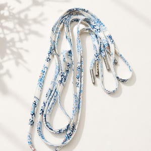 May include: A pair of flat shoelaces featuring a white base with a repeating blue and light brown ikat-style pattern. The laces are finished with metallic silver aglets at each end, providing a secure and polished look for footwear.