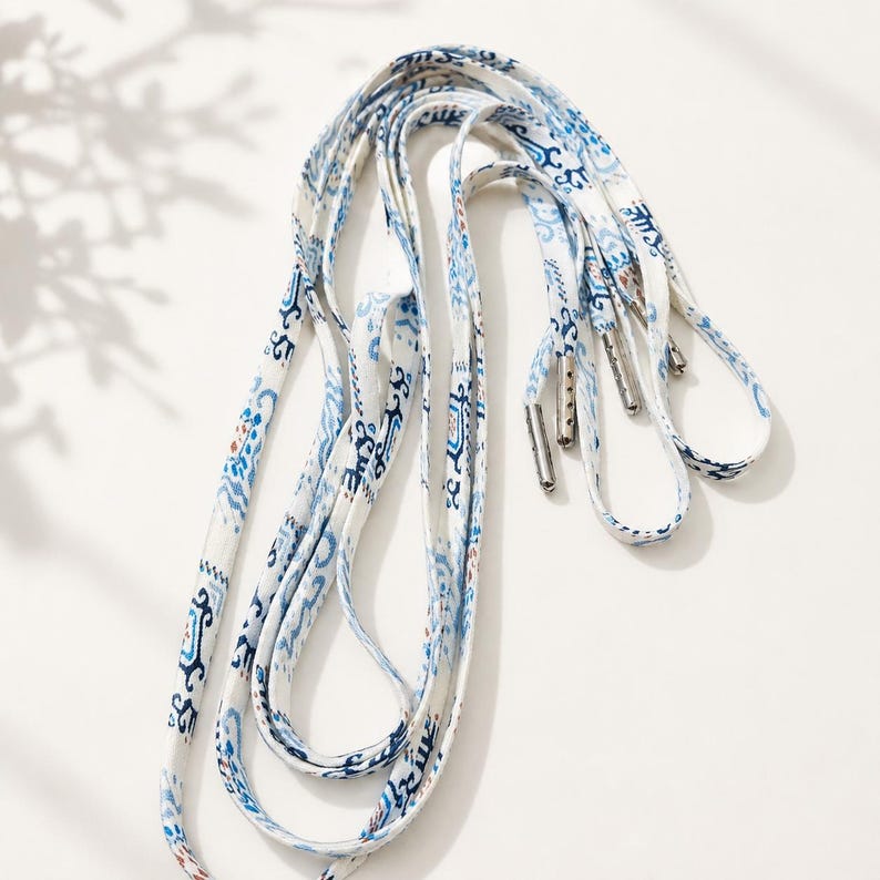 May include: A pair of flat shoelaces featuring a white base with a repeating blue and light brown ikat-style pattern. The laces are finished with metallic silver aglets at each end, providing a secure and polished look for footwear.
