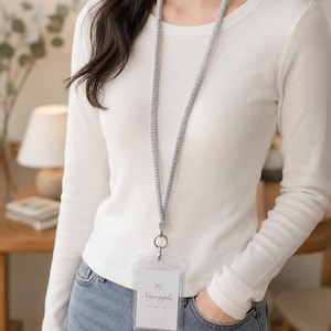 May include: A grey fabric lanyard with a silver metal clasp and a clear plastic vertical ID badge holder. The badge insert features a small bow icon above the text "Nanepple" and the phrase "MADE WITH LOVE" printed in black ink.