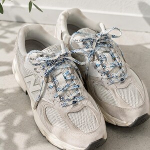May include: A pair of off-white New Balance sneakers featuring mesh and suede panels, a prominent N logo on the side, and patterned blue and white laces. The athletic shoes have a thick white midsole and a dark gray rubber outsole for traction.