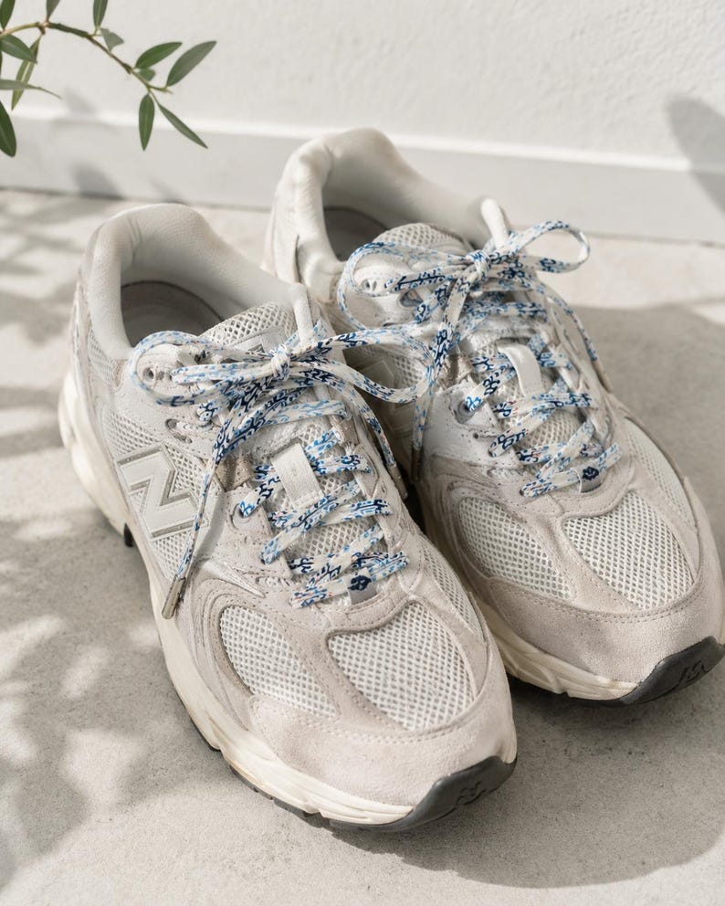 May include: A pair of off-white New Balance sneakers featuring mesh and suede panels, a prominent N logo on the side, and patterned blue and white laces. The athletic shoes have a thick white midsole and a dark gray rubber outsole for traction.