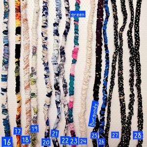 K&ouml;nnte beinhalten: Eine Sammlung von dreizehn langen, d&uuml;nnen Haaraccessoires aus Stoff, die vertikal angeordnet sind. Jedes St&uuml;ck hat eine geraffte Textur mit verschiedenen Mustern und Farben, darunter Blumen-, Punkte- und Unimuster, nummeriert von 16 bis 28.