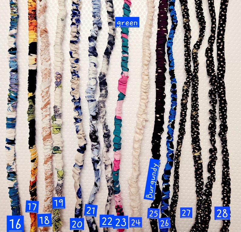 K&ouml;nnte beinhalten: Eine Sammlung von dreizehn langen, d&uuml;nnen Haaraccessoires aus Stoff, die vertikal angeordnet sind. Jedes St&uuml;ck hat eine geraffte Textur mit verschiedenen Mustern und Farben, darunter Blumen-, Punkte- und Unimuster, nummeriert von 16 bis 28.