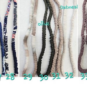 Op de afbeelding: Een collectie van elf met stof beklede elastische hoofdbanden gerangschikt in een rij, gelabeld met de nummers 25 tot en met 35. De banden hebben diverse patronen, waaronder bloemmotieven, abstracte prints en effen kleuren zoals olijf, haver, antraciet en blauw.