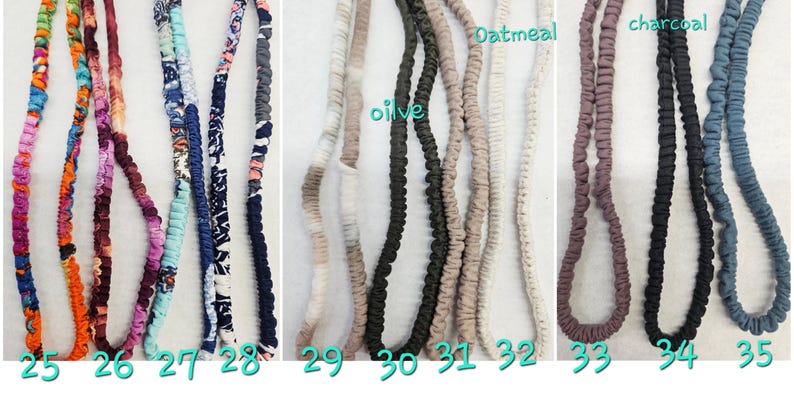 Op de afbeelding: Een collectie van elf met stof beklede elastische hoofdbanden gerangschikt in een rij, gelabeld met de nummers 25 tot en met 35. De banden hebben diverse patronen, waaronder bloemmotieven, abstracte prints en effen kleuren zoals olijf, haver, antraciet en blauw.