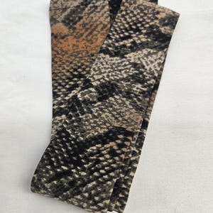 Puede incluir: Leggings estampados con dise&ntilde;o de piel de serpiente. Los leggings presentan una mezcla de colores negro, marr&oacute;n y beige, creando una apariencia realista de escamas de reptil. La tela est&aacute; doblada, mostrando el detalle y la textura del patr&oacute;n.