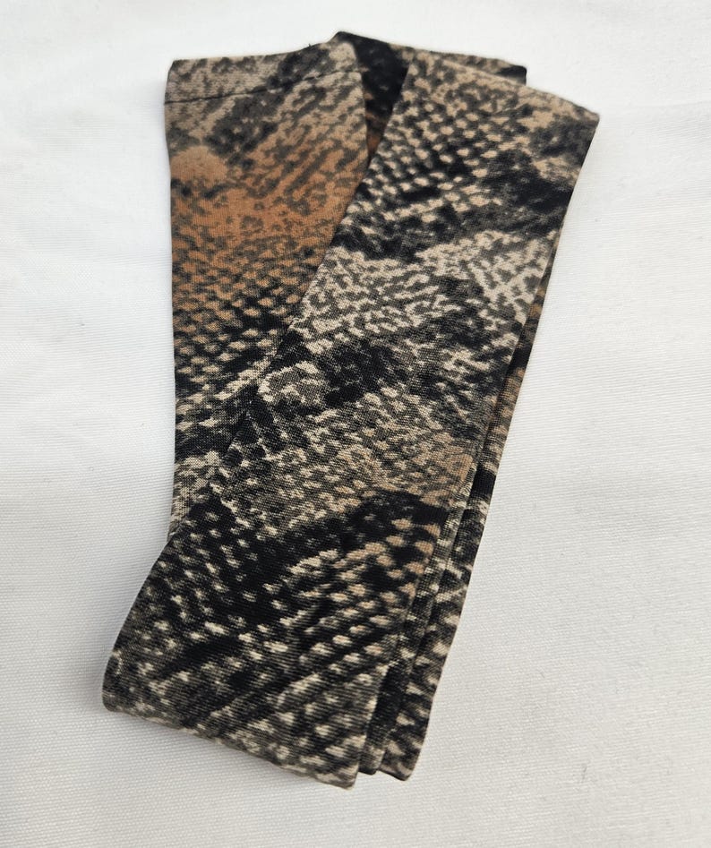 Puede incluir: Leggings estampados con dise&ntilde;o de piel de serpiente. Los leggings presentan una mezcla de colores negro, marr&oacute;n y beige, creando una apariencia realista de escamas de reptil. La tela est&aacute; doblada, mostrando el detalle y la textura del patr&oacute;n.