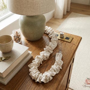 May include: A beige fabric-covered USB charging cable is draped across a wooden nightstand. A ceramic lamp with a beige shade sits atop a green ceramic base. A small ceramic cup and a jar with decorative items are also on the nightstand.