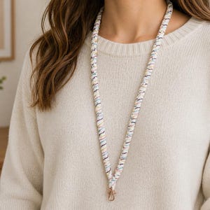 Op de afbeelding: Een stoffen lanyard met een gevlochten ontwerp, een witte basis en dunne, veelkleurige strepen in pasteltinten zoals blauw, roze en geel. De lanyard is voorzien van een goudkleurige metalen karabijnhaak aan de onderkant.