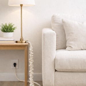 Puede incluir: Una lámpara dorada con una pantalla blanca se encuentra sobre una mesa auxiliar de madera junto a un sofá crema. El cable de la lámpara está cubierto por una funda de tela color crema. El texto "Cozy cords, clean look" es visible.