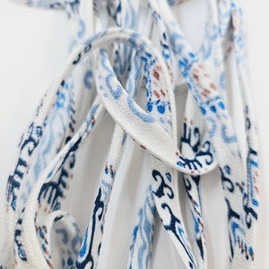 May include: A long, thin fabric ribbon or shoelace featuring a white base color with a repeating pattern of abstract shapes and lines in shades of dark blue, light blue, and brown. The material has a woven texture and is coiled in a loose pile.